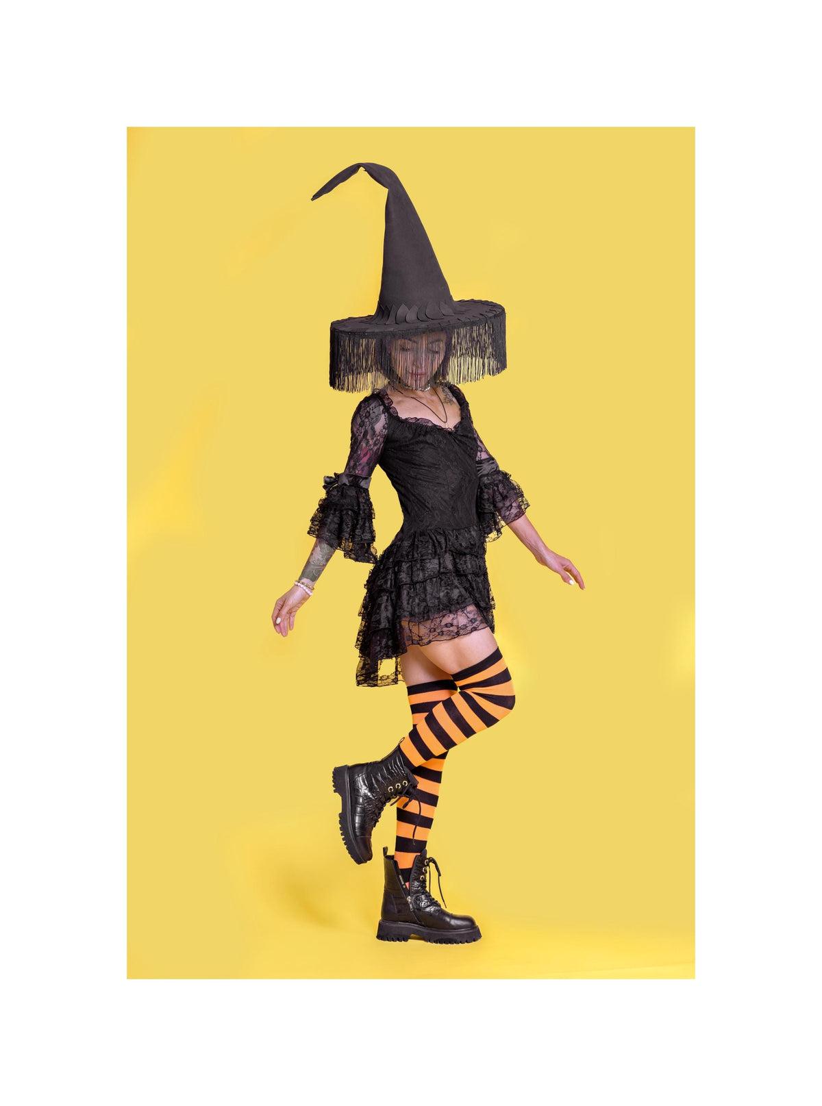 May include: A black witch costume with a tall, pointed hat, a lace dress, and striped orange and black thigh-high socks. The costume includes black boots. The background is a solid yellow.