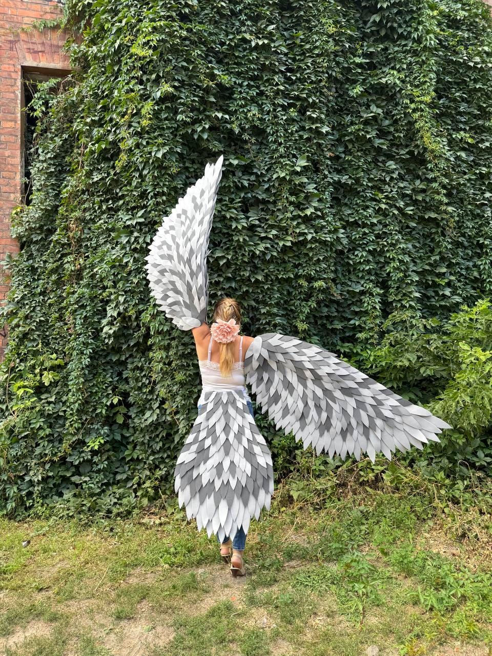 May include: A costume with large, feathered wings in shades of gray and white. The wings are attached to a matching skirt, creating a bird-like appearance. The costume is worn outdoors against a backdrop of green foliage.