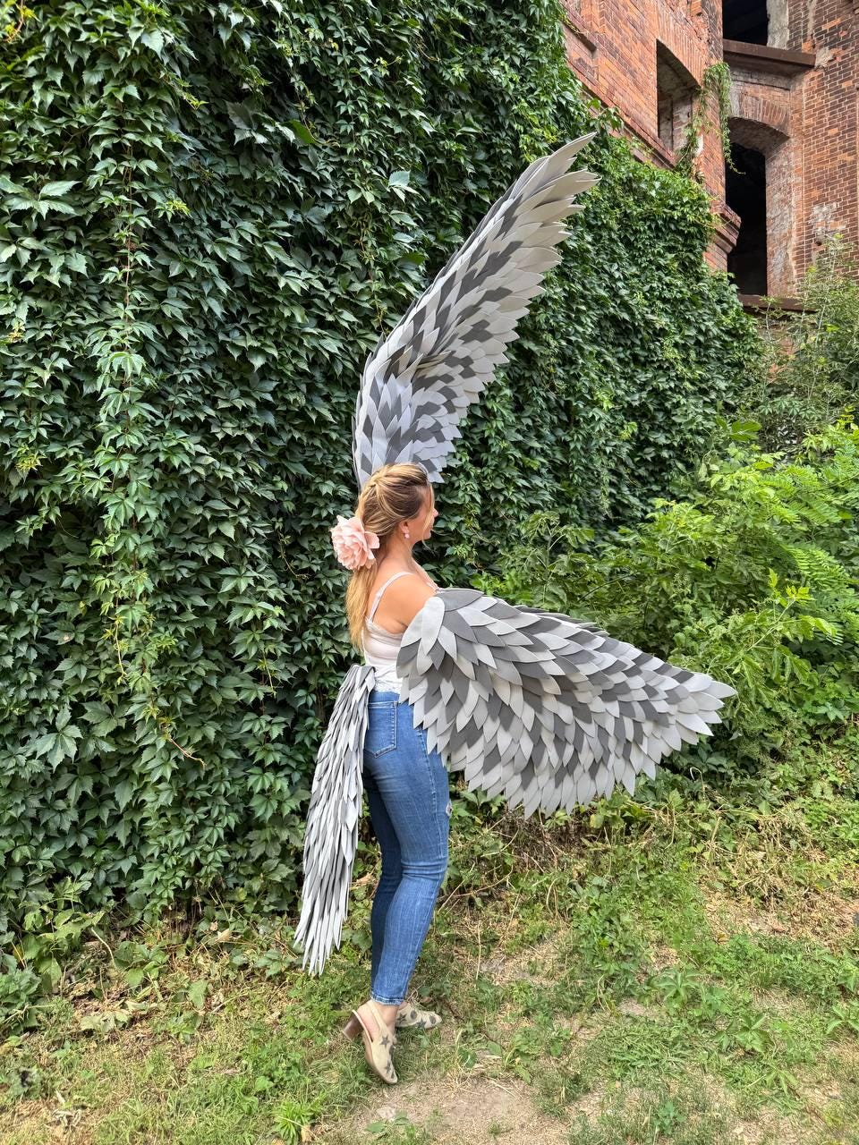 May include: A person wearing large, gray and white feathered wings and a matching tail. The wings are layered with a gradient of gray and white feathers. The person is wearing a white top and blue jeans, standing in front of a wall of green foliage.