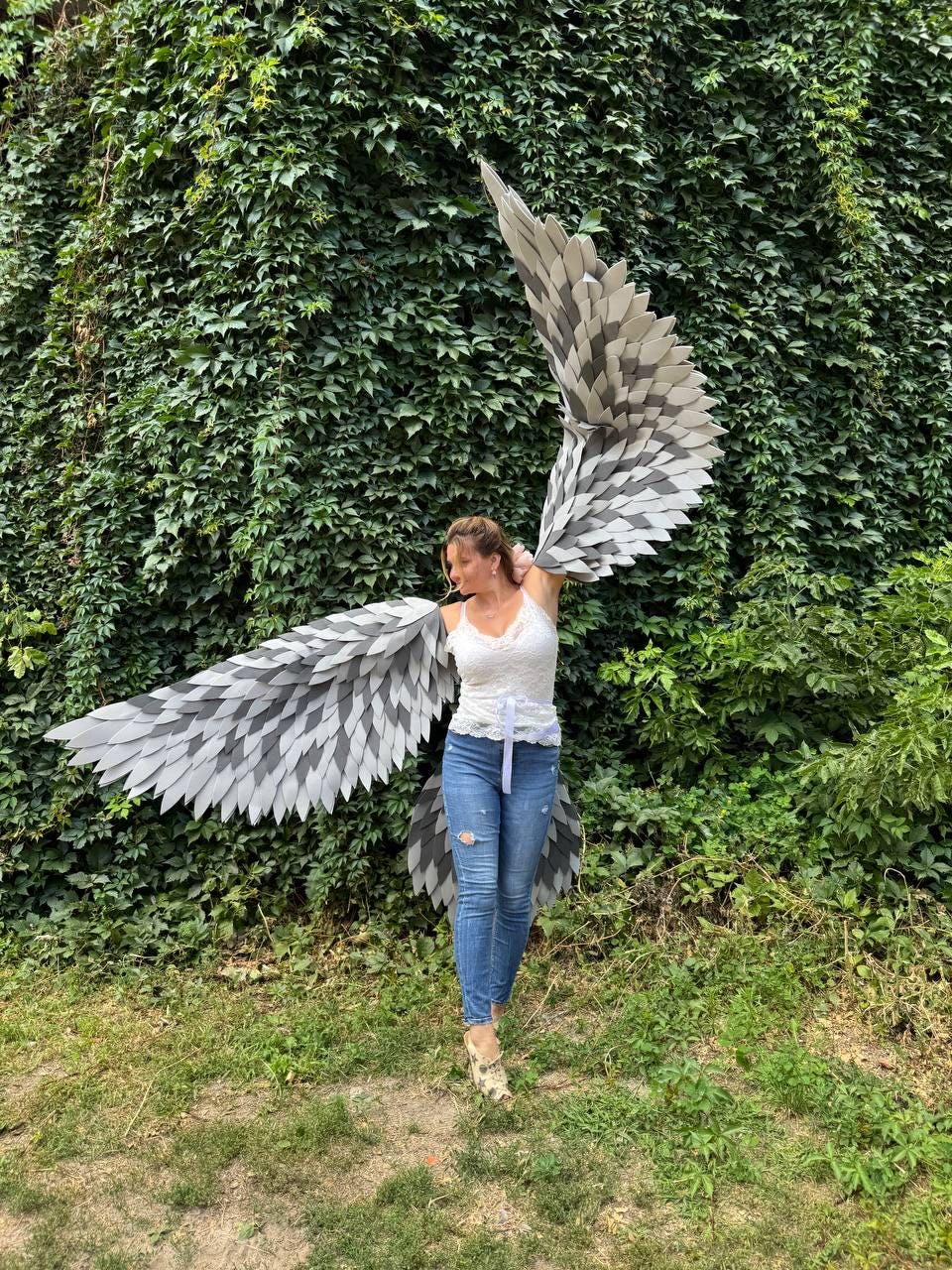 May include: A person wearing large, gray and white feathered wings, standing in front of a wall of green foliage. The wings are spread wide, showcasing a layered design with a gradient of colors. The person is wearing a white top and blue jeans.