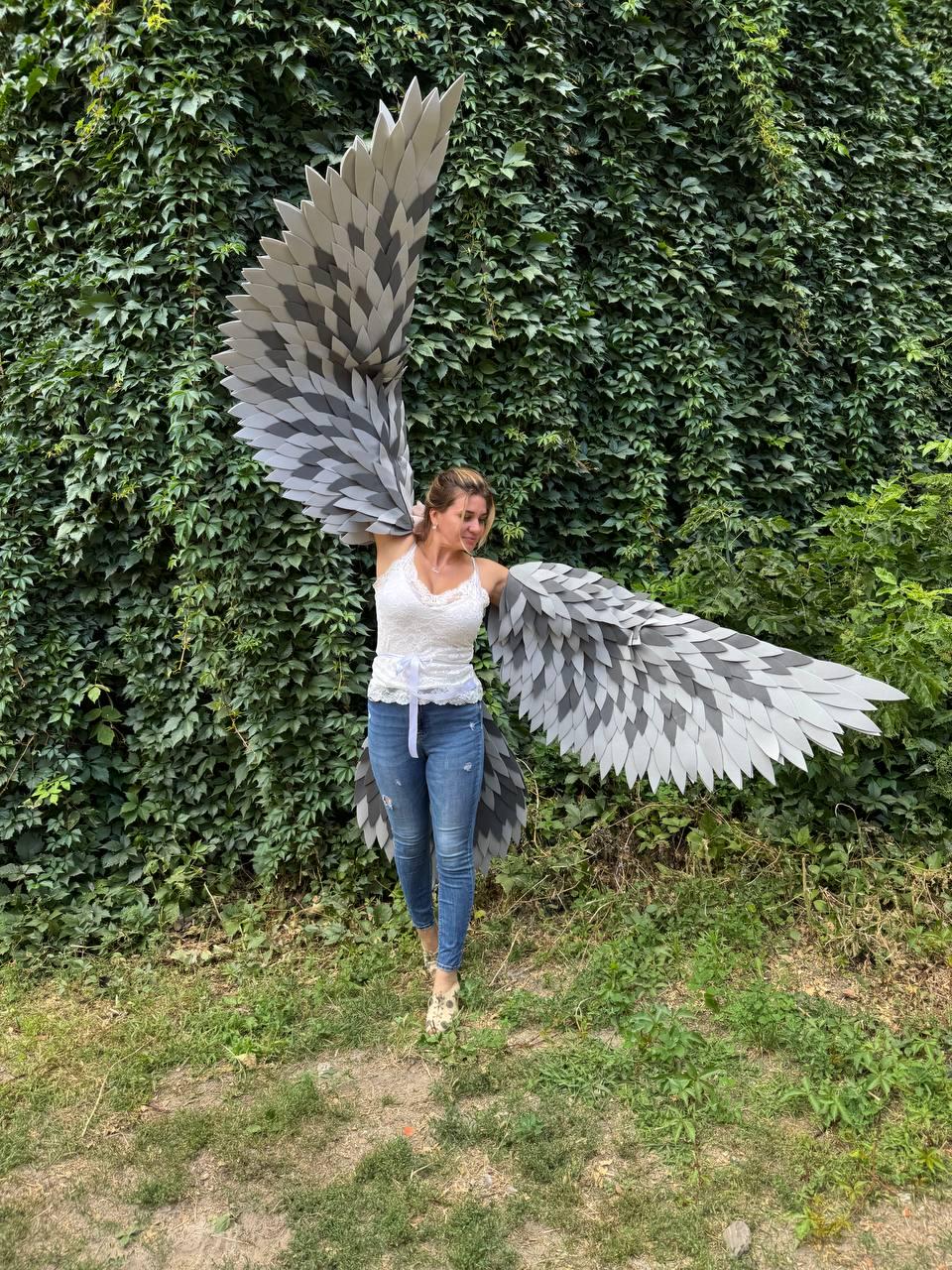 May include: Gray and white feathered wings with a layered design. The wings are worn by a person wearing a white top and blue jeans. The wings are spread wide against a backdrop of green foliage.