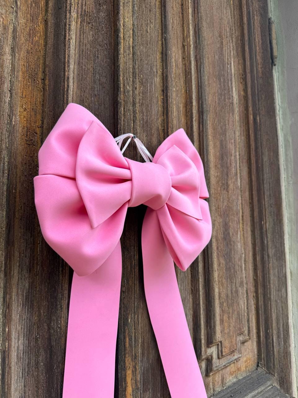 May include: A large, bright pink bow with long, flowing ribbons. The bow is made of a smooth, satin-like material and is suspended against a weathered, wooden door. The bow's color is a vibrant, eye-catching shade of pink.
