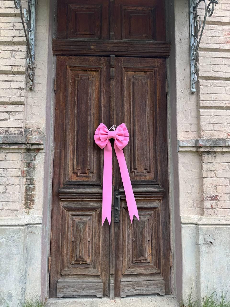 May include: A large, bright pink bow with long streamers is attached to a weathered, dark brown wooden door. The door has a paneled design and is set in a brick and stone facade.
