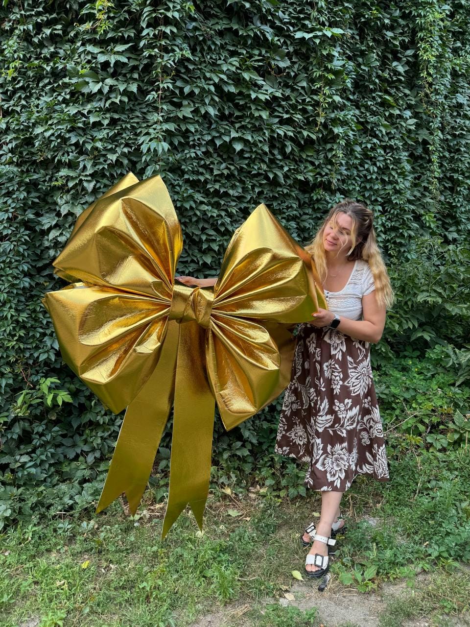 May include: A large, golden bow with long, flowing ribbons. The bow is made of a shiny, metallic material and is positioned in front of a wall of green foliage. The bow is a decorative element, suitable for events or celebrations.