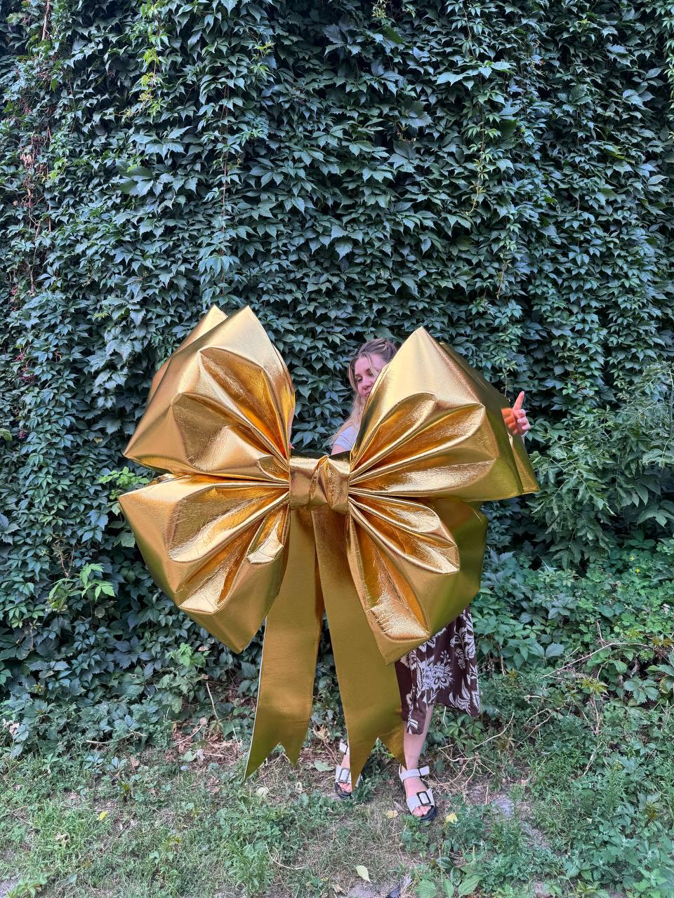 May include: A large, shiny gold bow with long, flowing ribbons. The bow is made of a metallic material and is positioned in front of a wall of green foliage. The bow is a decorative element, possibly for a gift or event.