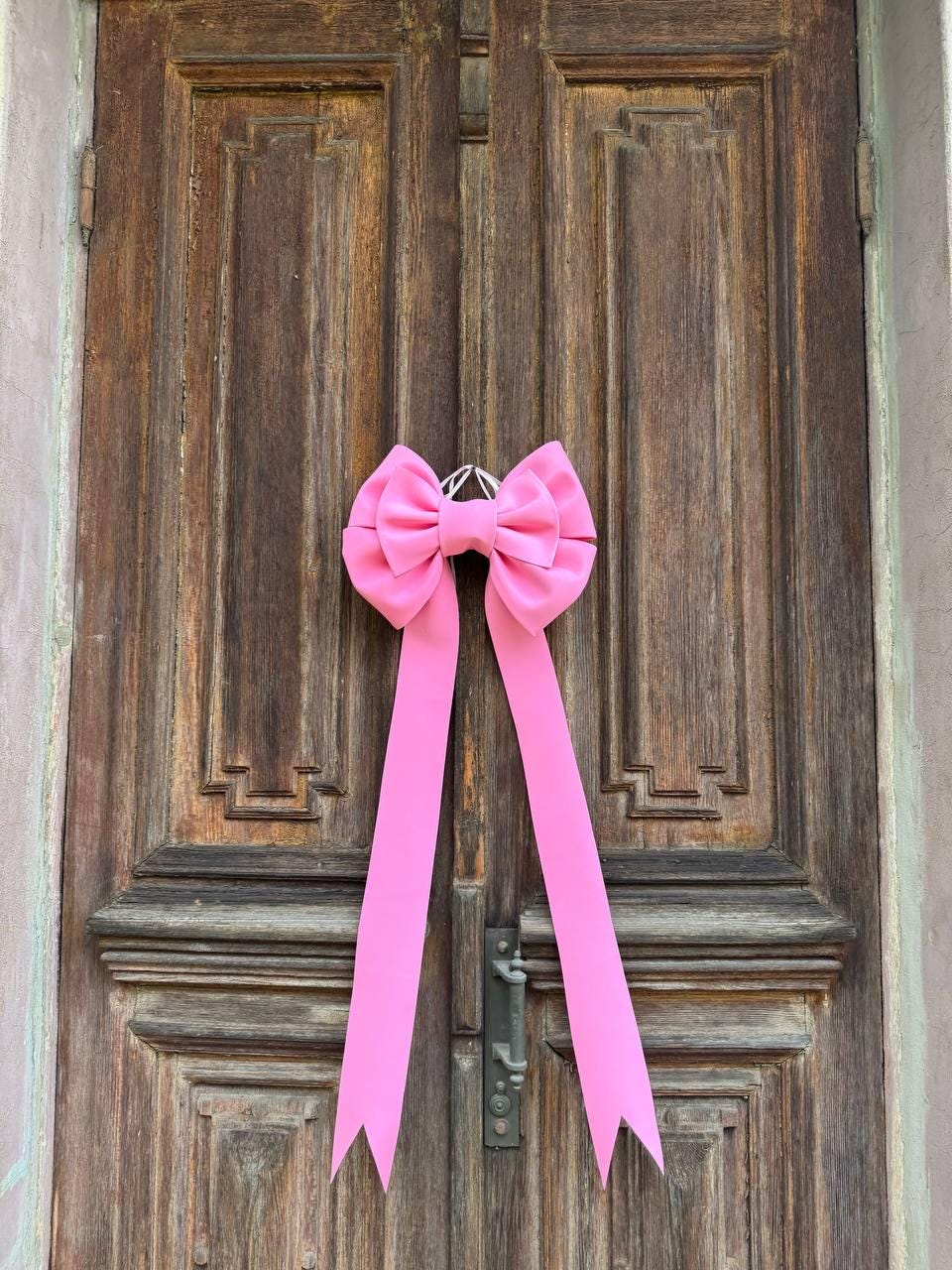May include: A bright pink bow with long streamers hangs on a weathered wooden door. The bow is made of a soft, possibly satin-like material, and is attached to the door with a silver-colored metal ring. The door has a dark, aged appearance.