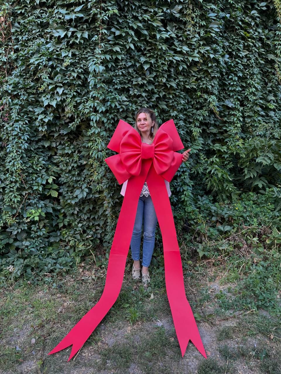 May include: A large, vibrant red bow with long, flowing ribbons. The bow is held in front of a person standing in front of a wall of green foliage. The bow's dimensions are substantial, making it a striking decorative element.