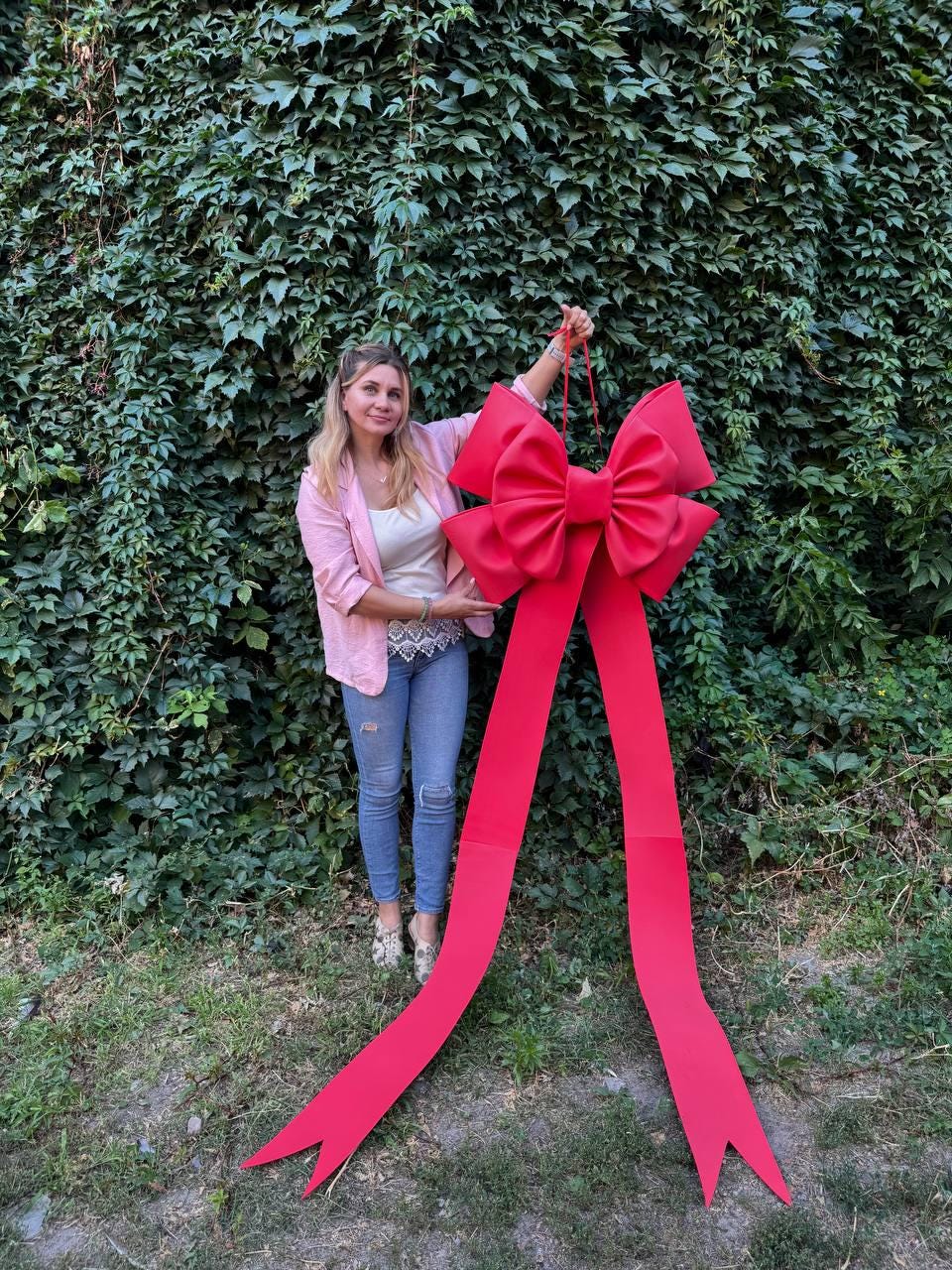 May include: A large, bright red bow with long, flowing ribbons. The bow is held up against a backdrop of lush green foliage. The bow is a festive decoration, perfect for gifts or events.