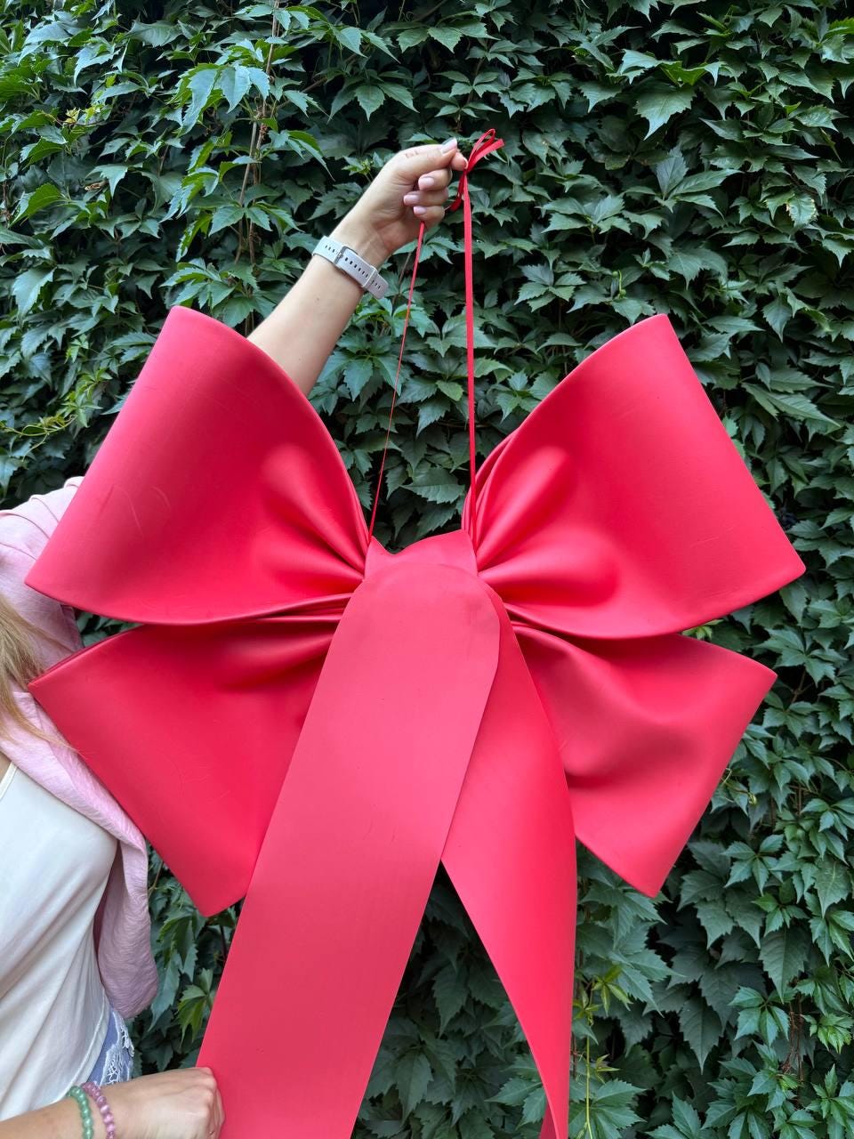 May include: A large, vibrant red bow with long, flowing ribbons. The bow is held up against a backdrop of green foliage. The bow's size and color make it suitable for festive decorations.