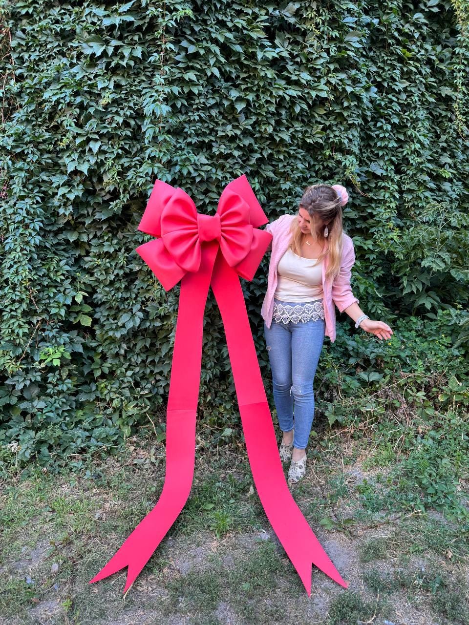 May include: A large, vibrant red bow with long, flowing ribbons. The bow is made of a smooth material and is positioned in front of a lush green wall of foliage. The bow's ribbons extend downwards, creating a striking visual contrast.