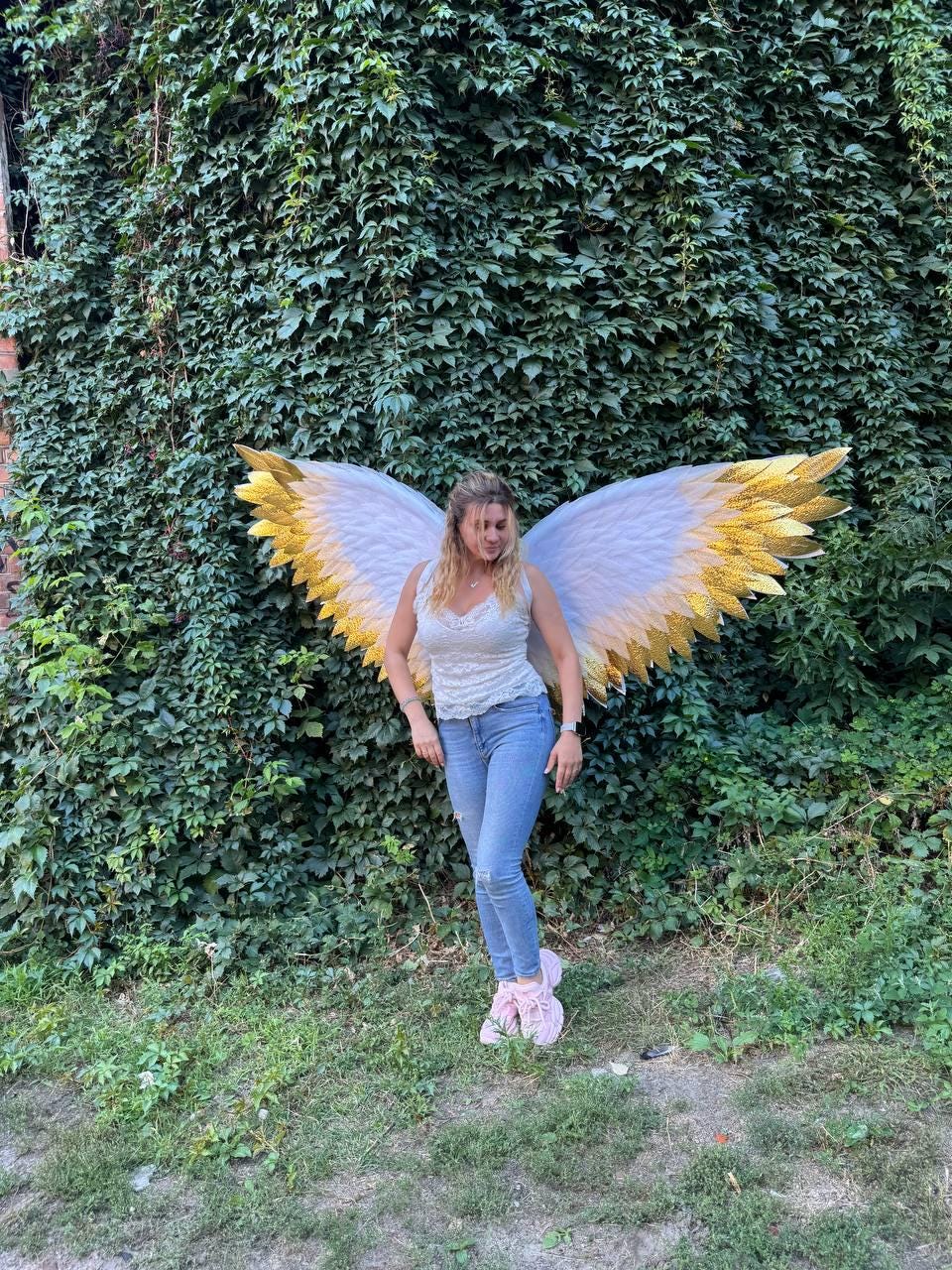 May include: A person wearing large angel wings with white and gold feathers stands in front of a wall of green foliage. The wings are the focal point, with their detailed design and contrasting colors. The person is wearing a white top, blue jeans, and pink shoes.