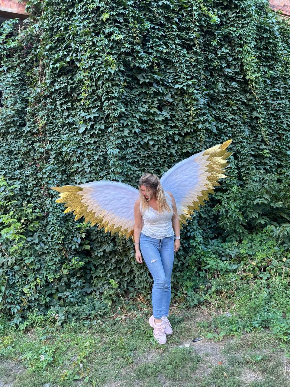 May include: Large, decorative angel wings with white and gold feathers. The wings are displayed against a backdrop of lush green foliage. The wings are a costume accessory.