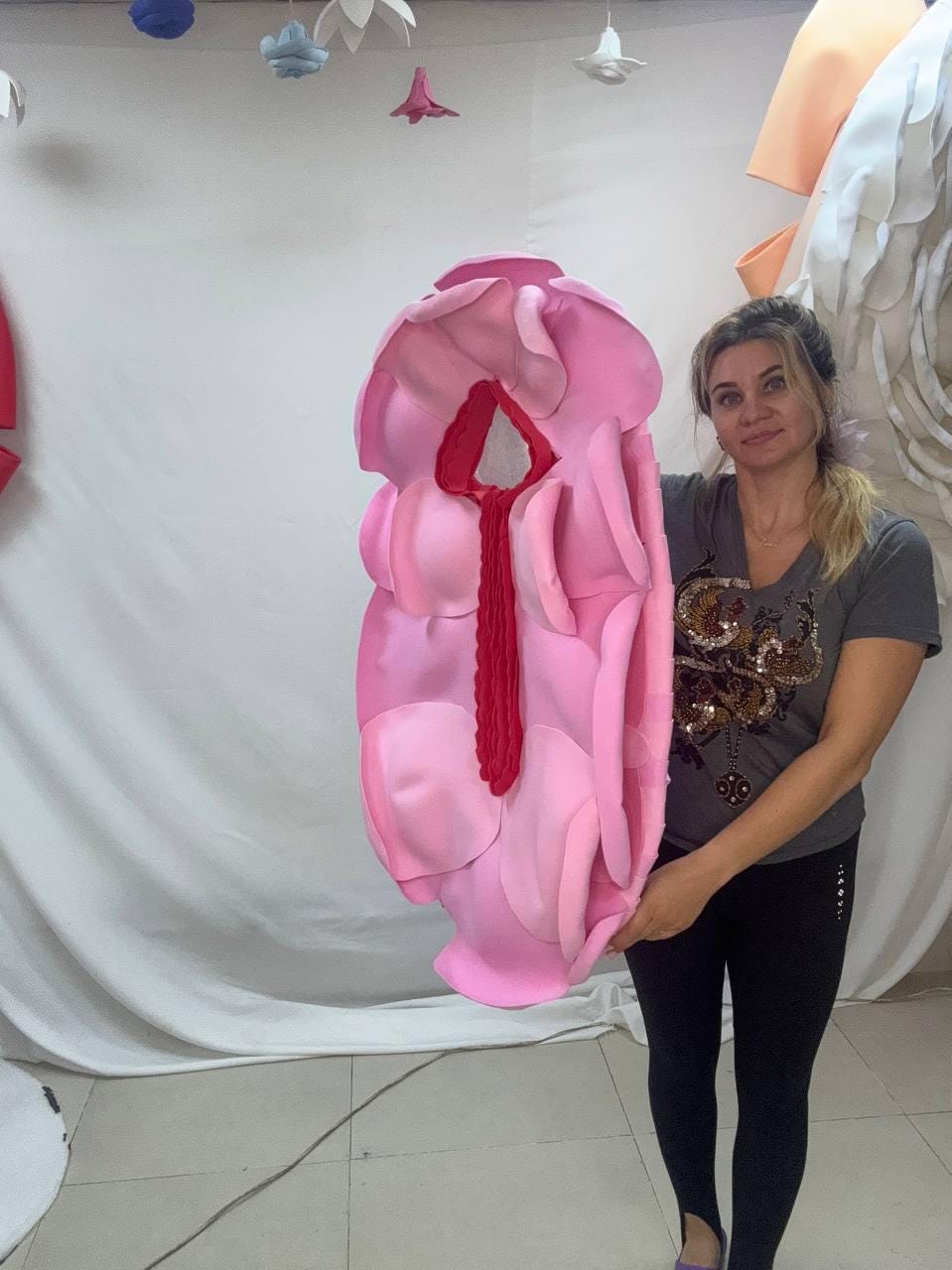 May include: A large, pink, flower-shaped sculpture with a red center. The sculpture is held by a person. The sculpture is made of soft, layered material, with varying shades of pink. The background is a white wall with hanging paper decorations.