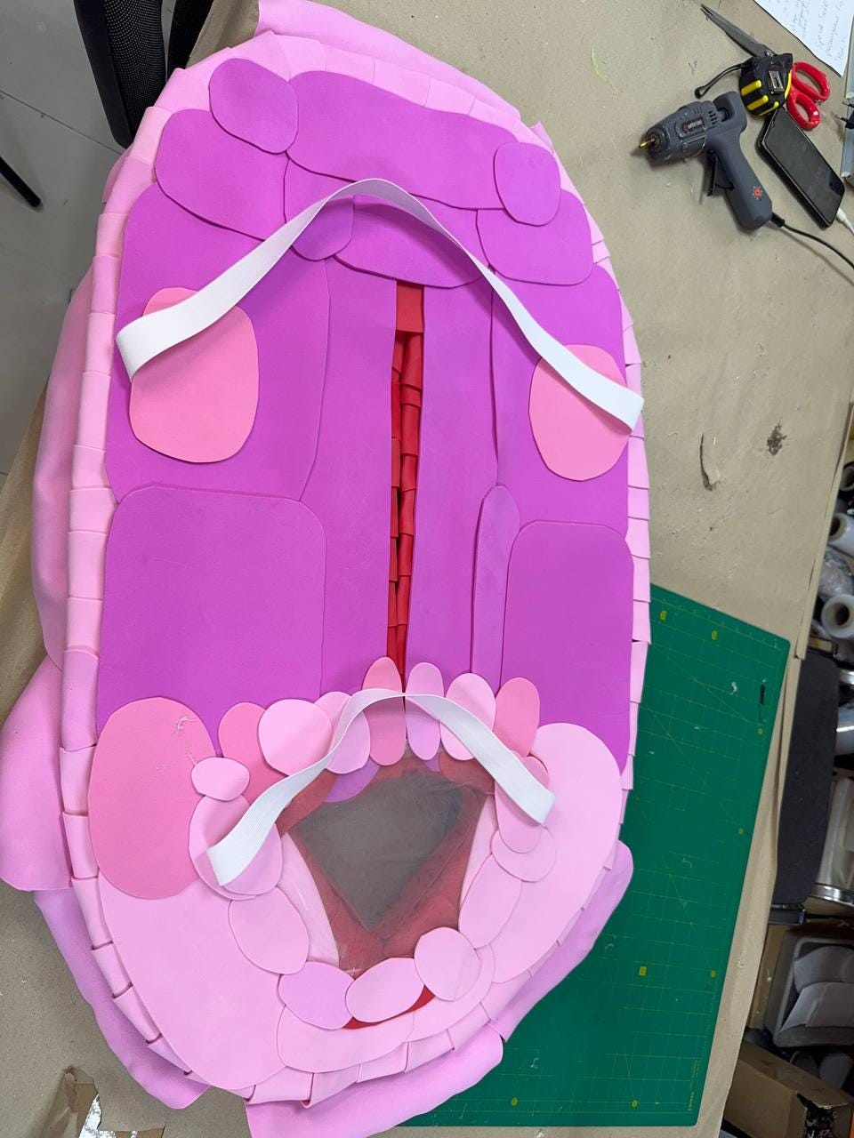 May include: A handmade, pink and purple foam turtle shell with white straps. The shell is constructed from layered foam pieces, with a red center and a dark gray area. Crafting tools, including a glue gun and scissors, are visible in the background.