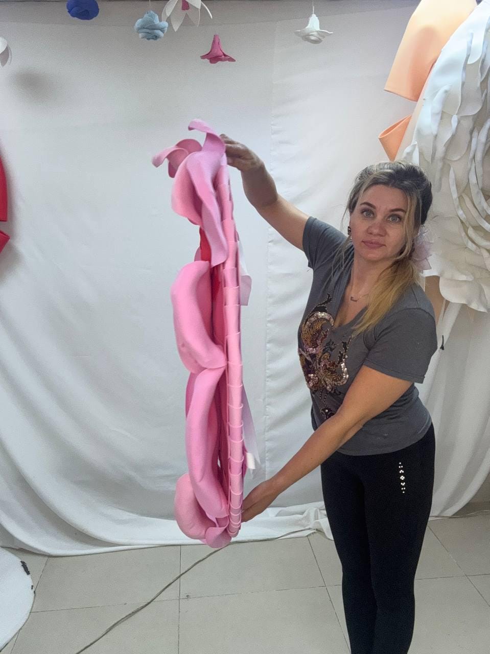 May include: A large, pink, layered flower decoration is held up. The flower has multiple petals and a structural frame. The background includes other paper flower decorations and a white curtain.