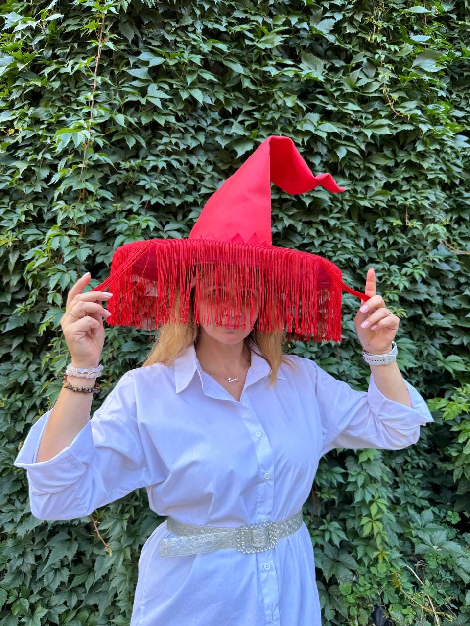 May include: A bright red witch hat with a pointed crown and a wide brim adorned with long, flowing fringe. The hat is the focal point, suggesting a Halloween or costume theme. The hat is worn by a person wearing a white dress.