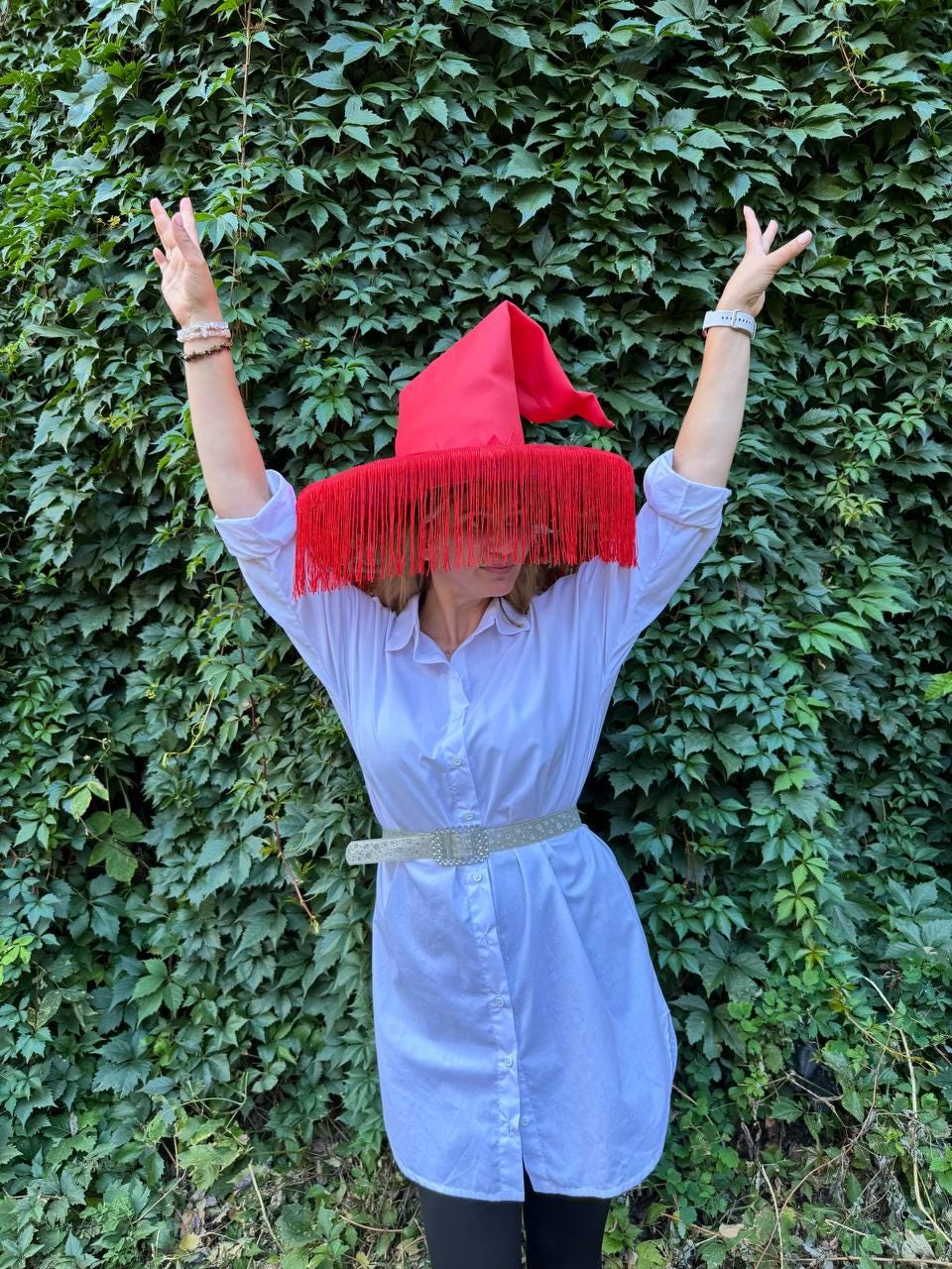 May include: A red witch hat with long fringe, worn with a white button-down shirt and a silver belt. The hat's pointed crown and fringe obscure the face. The background is a wall of green leaves.