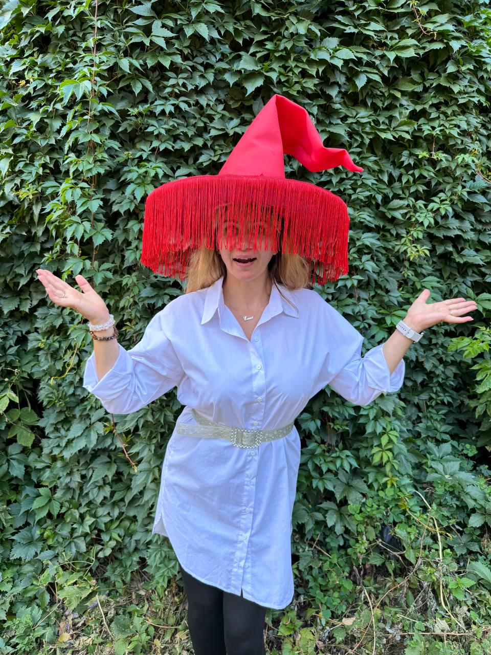 May include: A red witch hat with long fringe, worn over a white button-down shirt and a silver belt. The hat's pointed crown and fringe create a dramatic effect. The background is a wall of green leaves.
