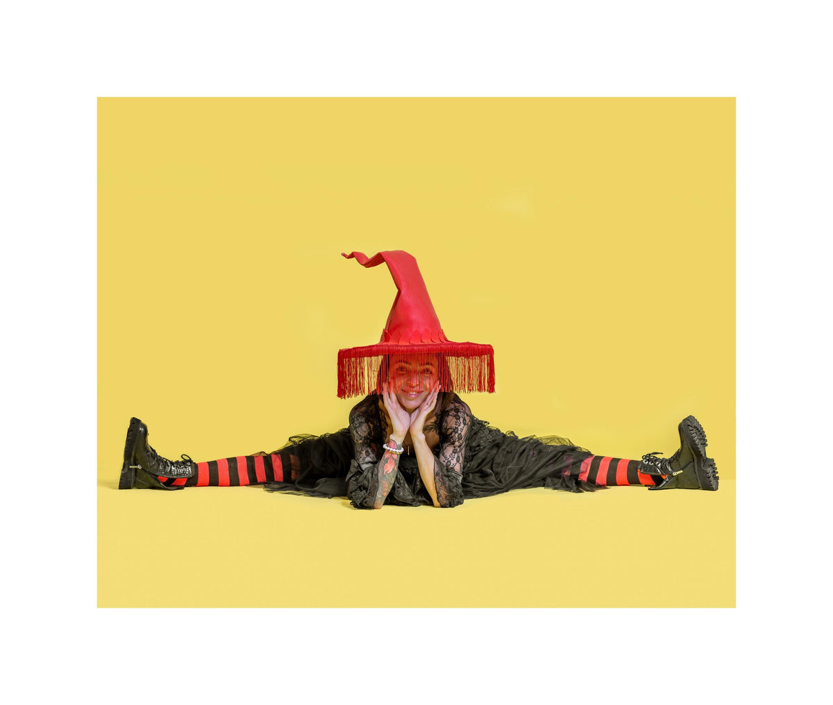 May include: A person wearing a red witch hat with a fringed brim, black boots, and black and red striped socks. The person is wearing a black dress and is lying on a yellow background.