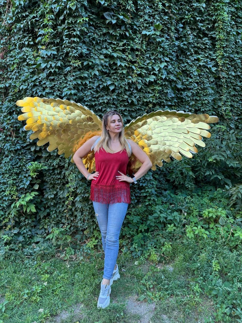 May include: A woman wearing a red top and blue jeans stands in front of a wall of green foliage, with large, golden angel wings attached to her back. The wings are detailed with layered feathers.