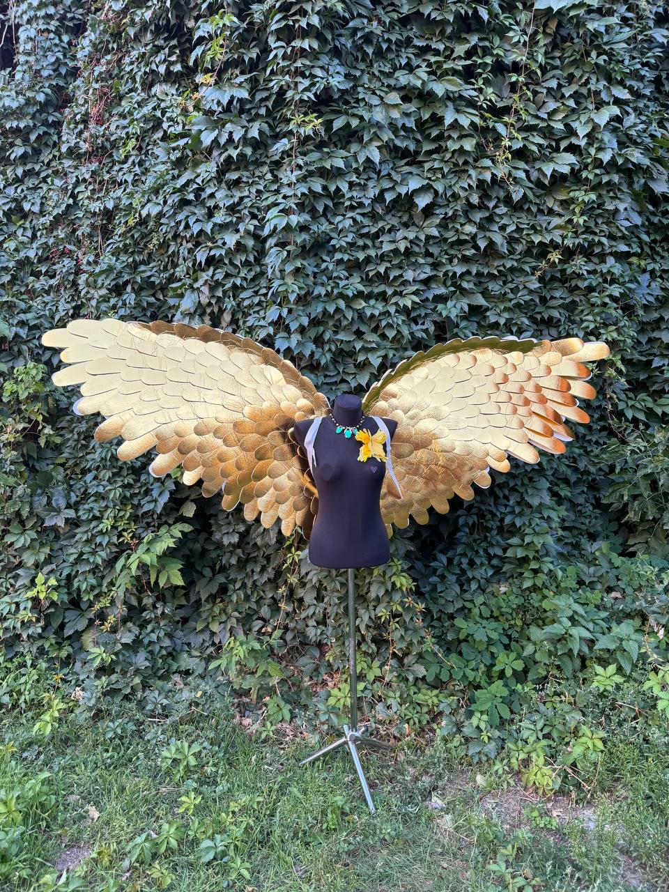 May include: A pair of large, golden-colored angel wings displayed on a black mannequin. The wings have a layered design with a gradient of colors from gold to brown. A necklace with turquoise beads and a yellow flower adorn the mannequin.