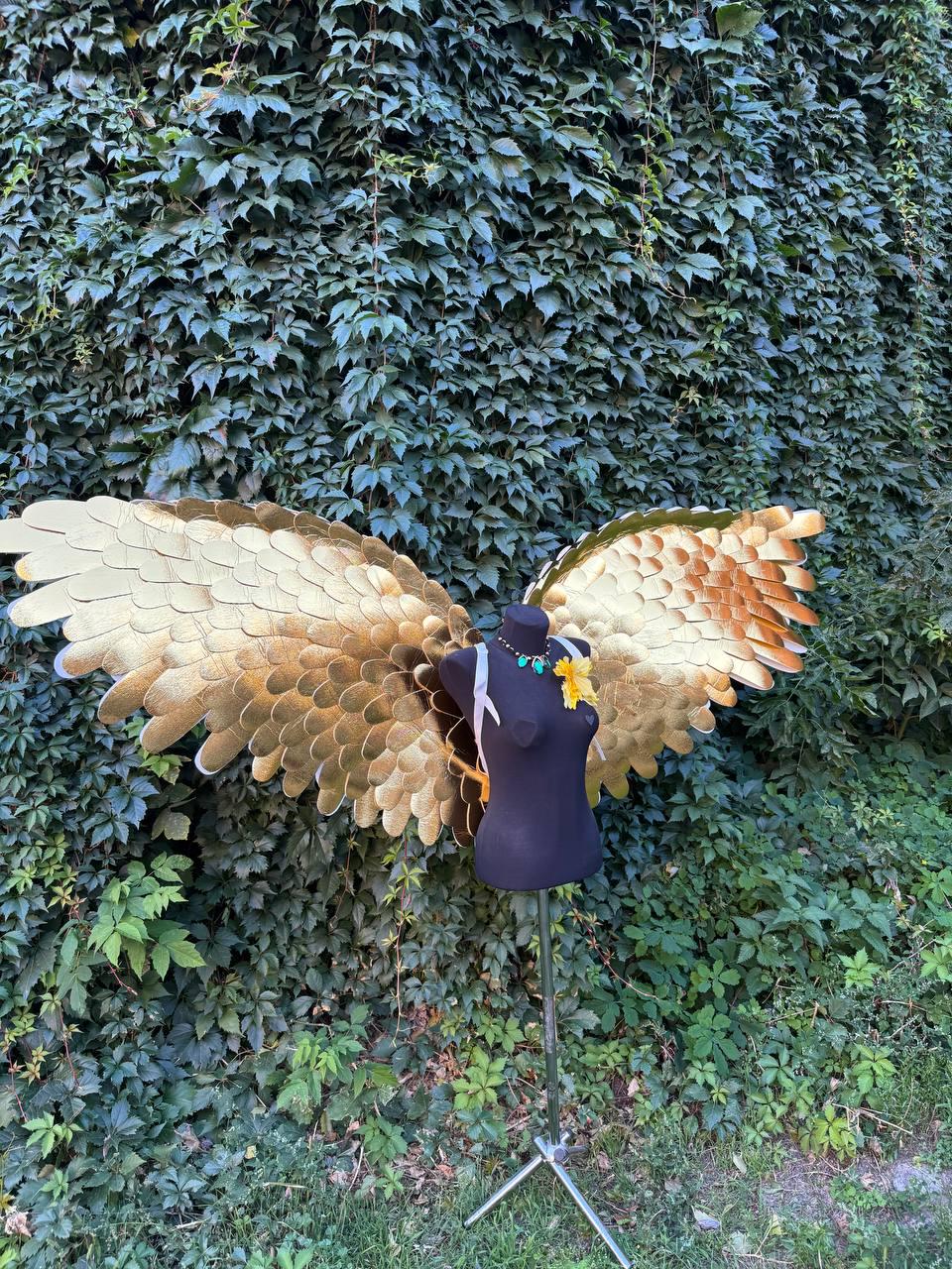 May include: A pair of large, golden-colored angel wings with detailed feathering, displayed on a black mannequin. The wings are set against a backdrop of lush green foliage, suggesting an outdoor setting. A necklace and a yellow flower adorn the mannequin.