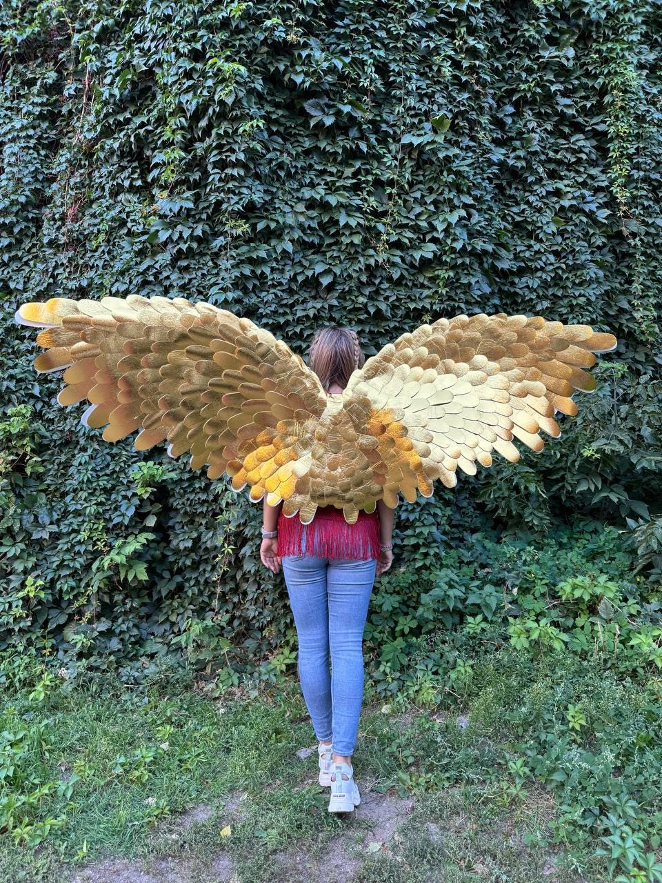 May include: A person wearing large, golden-colored angel wings with a layered feather design. The wings are the focal point, set against a backdrop of lush green foliage. The person is wearing a red top and blue jeans.