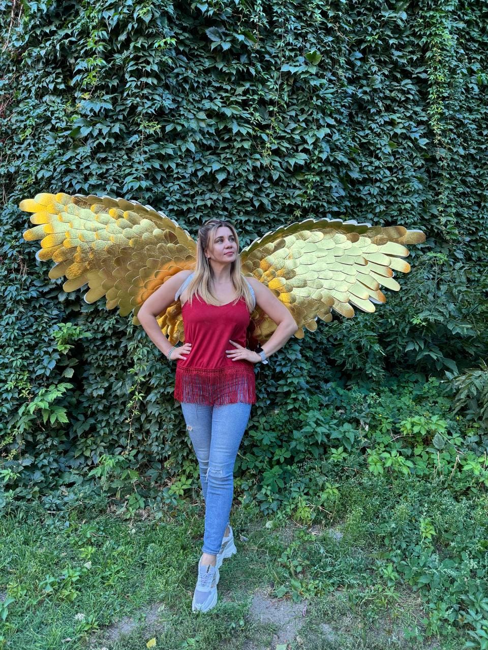 May include: A woman wearing gold-colored angel wings stands in front of a wall of green foliage. She is wearing a red top, blue jeans, and gray sneakers. The wings are large and detailed, with a layered feather design.