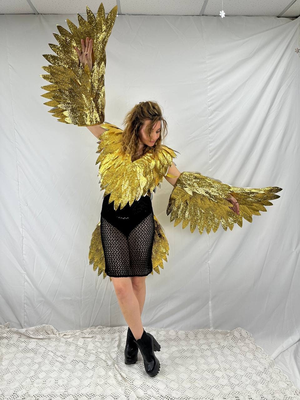 May include: A woman wearing a gold sequined bird costume with large, outstretched wings. The costume includes a matching collar and arm pieces, paired with a black dress and boots. The wings are a prominent feature, catching the light.