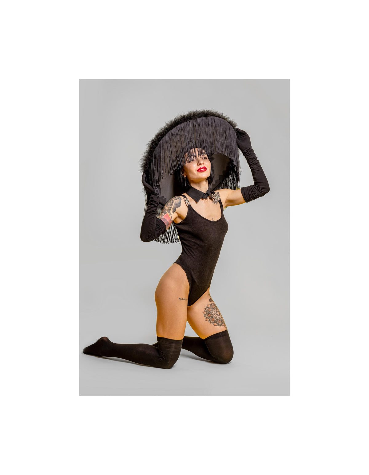 May include: A black bodysuit, knee-high socks, and long gloves are worn with a large black hat with fringe. The hat is the focal point, with the model kneeling on a gray background.