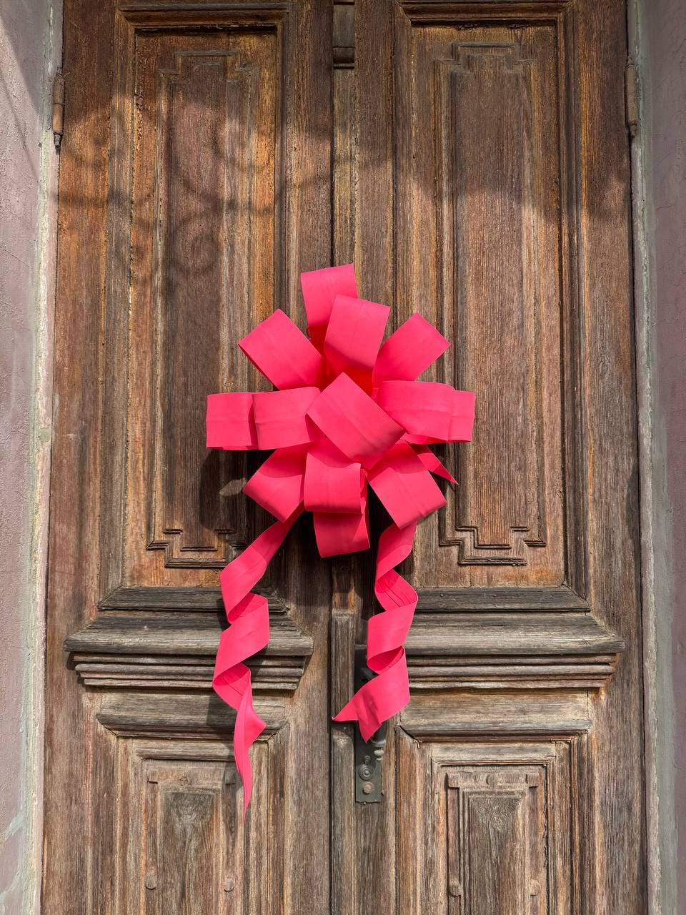 May include: A large, bright pink bow adorns a weathered wooden door. The bow is made of wide ribbon, with long, curling streamers. The door has a detailed, carved design and a dark metal handle.