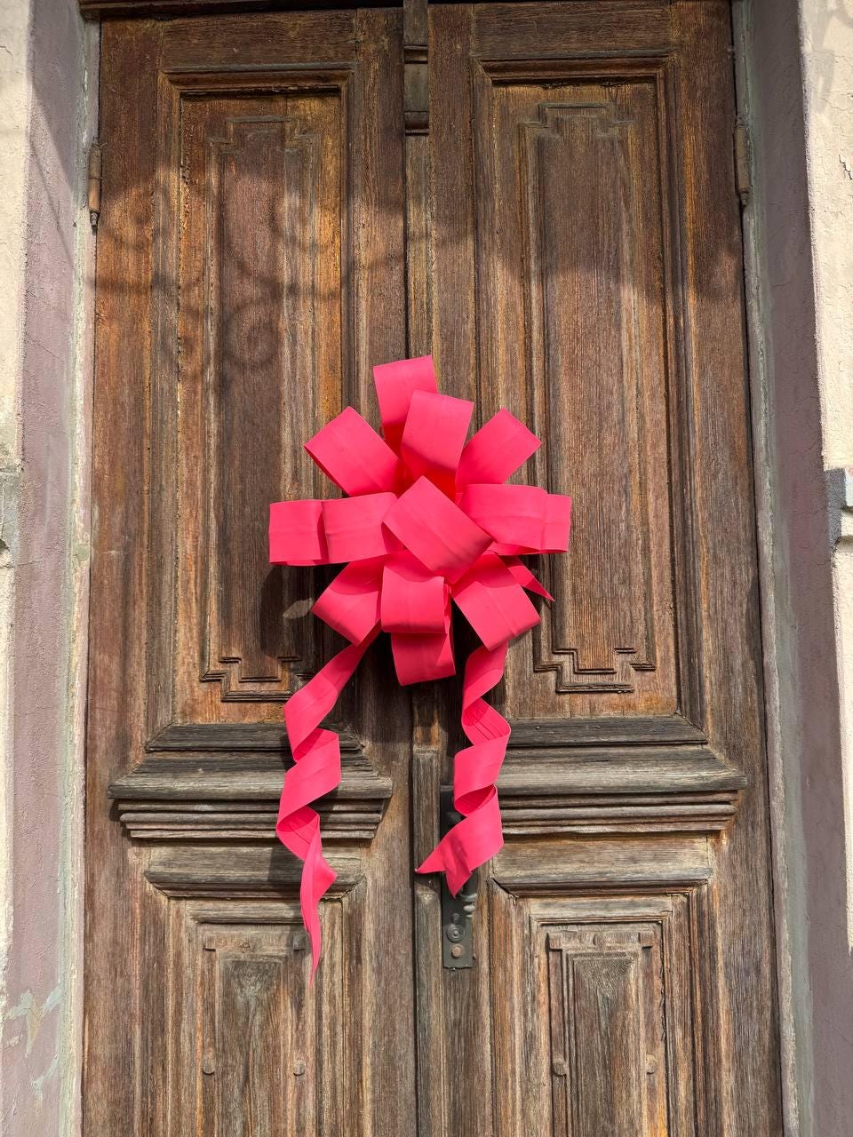 May include: A bright pink bow adorns a weathered wooden door. The bow is large and elaborate, with multiple loops and long, flowing ribbons. The door has a detailed, paneled design and a warm, brown color.