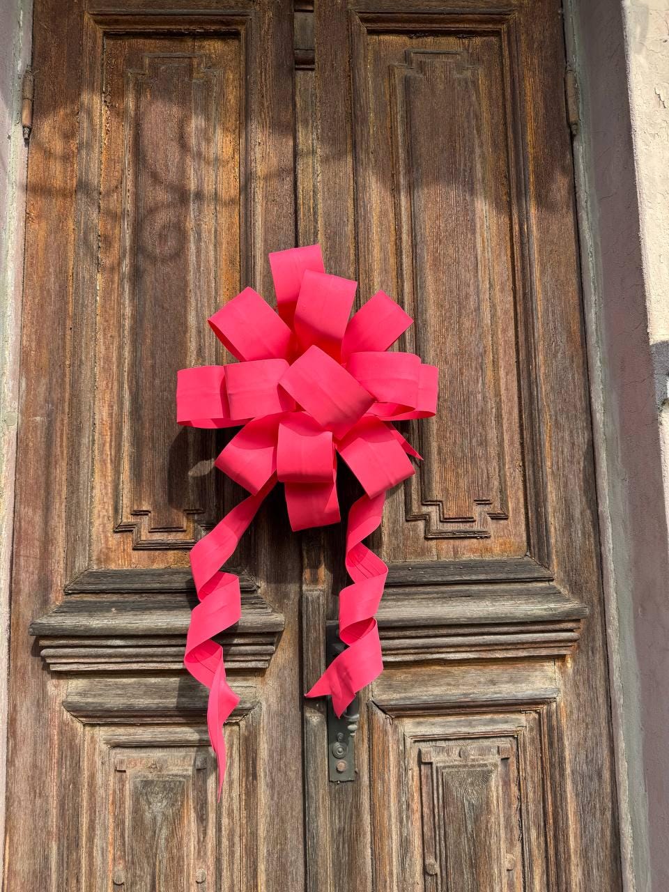 May include: A large, vibrant pink bow adorns a weathered wooden door. The bow's ribbons cascade downwards, adding a touch of elegance. The door's aged wood grain and intricate paneling provide a rustic backdrop to the bright, festive decoration.