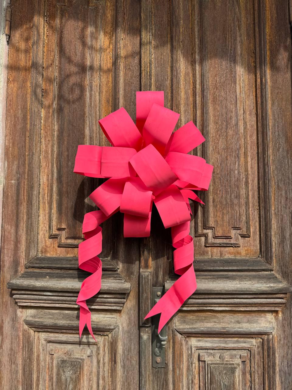 May include: A vibrant pink bow adorns a weathered wooden door. The bow's loops and cascading ribbons create a festive display. The door's aged wood grain and decorative panels provide a rustic backdrop for the bright accent.