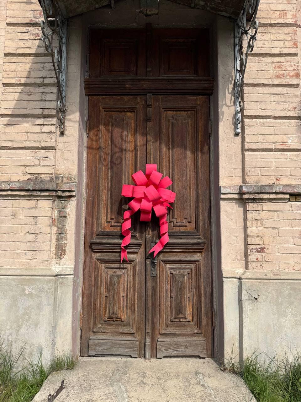 May include: A weathered, dark brown wooden door with intricate paneling. A large, bright pink bow is centered on the door. The door is set in a brick and concrete facade, with a decorative metal awning above.