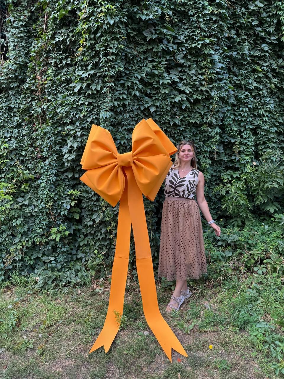 May include: A large, bright orange bow with long, flowing ribbons. The bow is made of a fabric material and stands tall against a backdrop of lush green foliage. The bow is a decorative element, possibly for an event or celebration.
