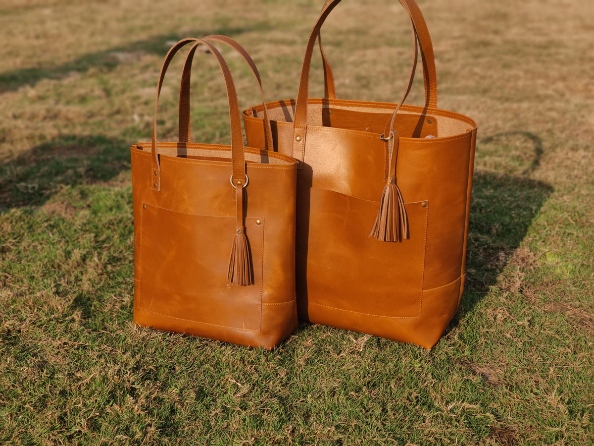 May include: Two brown leather tote bags of different sizes, standing on grass. Each bag has a front pocket, long handles, and a decorative tassel. The bags are a warm, medium brown color.