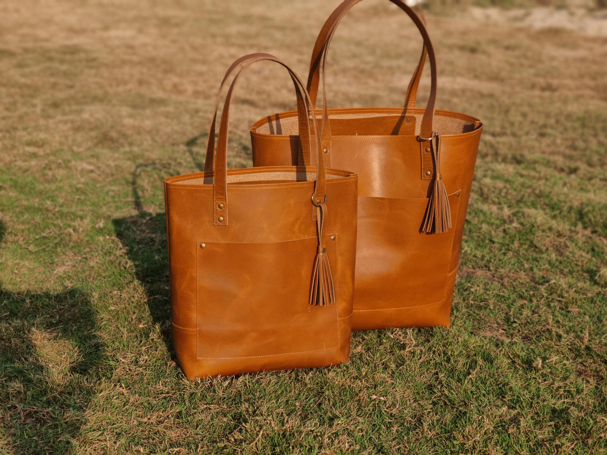 May include: Two brown leather tote bags of different sizes, with matching handles and exterior pockets. Each bag has a decorative tassel. The bags are set on a grassy surface.