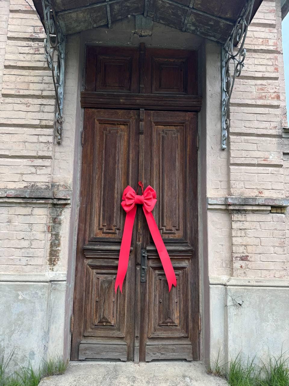 May include: A weathered, dark brown wooden door with intricate paneling. A bright red bow with long streamers is tied to the door. The door is set in a light pink brick building with a decorative metal awning.
