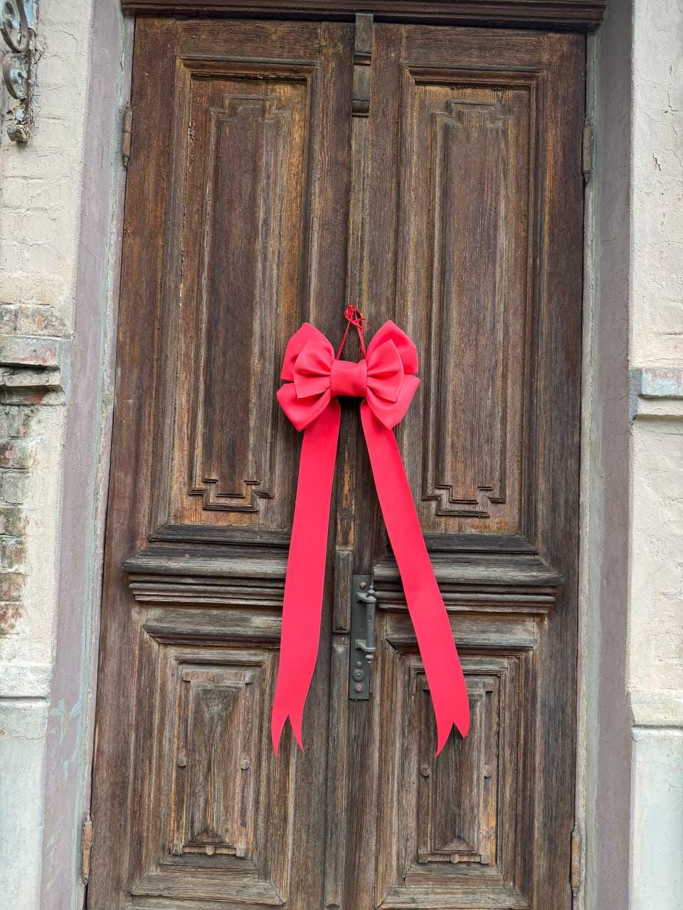 May include: A large, bright red bow with long streamers hangs on a weathered, wooden door. The door has a dark brown finish with detailed paneling and a metal handle. The bow is the focal point, adding a pop of color to the rustic door.