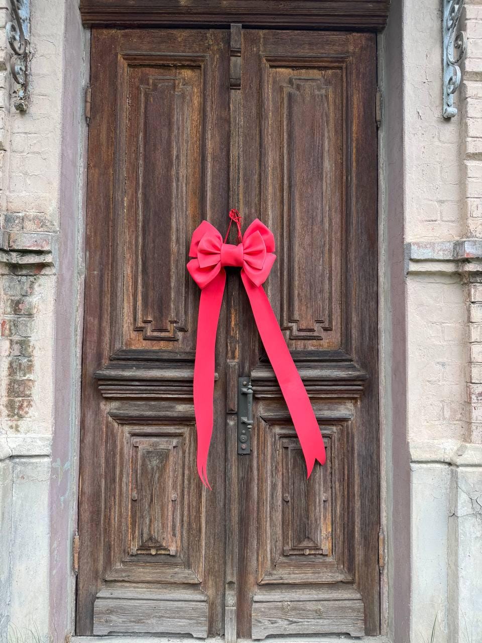May include: A bright red bow with long streamers hangs on a weathered, wooden door. The door has a detailed, paneled design and a metal handle. The surrounding wall is a light beige color.