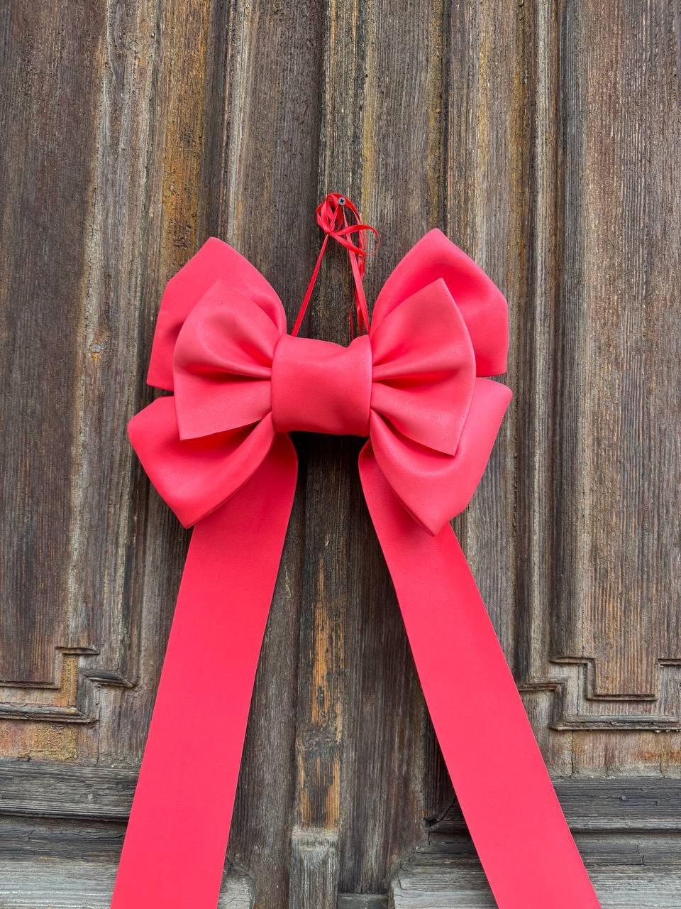 May include: A large, vibrant coral-colored bow with long, flowing ribbons hangs against a weathered wooden door. The bow is made of a smooth, satin-like material, and the ribbons cascade downwards. The bow is tied with a matching coral string.