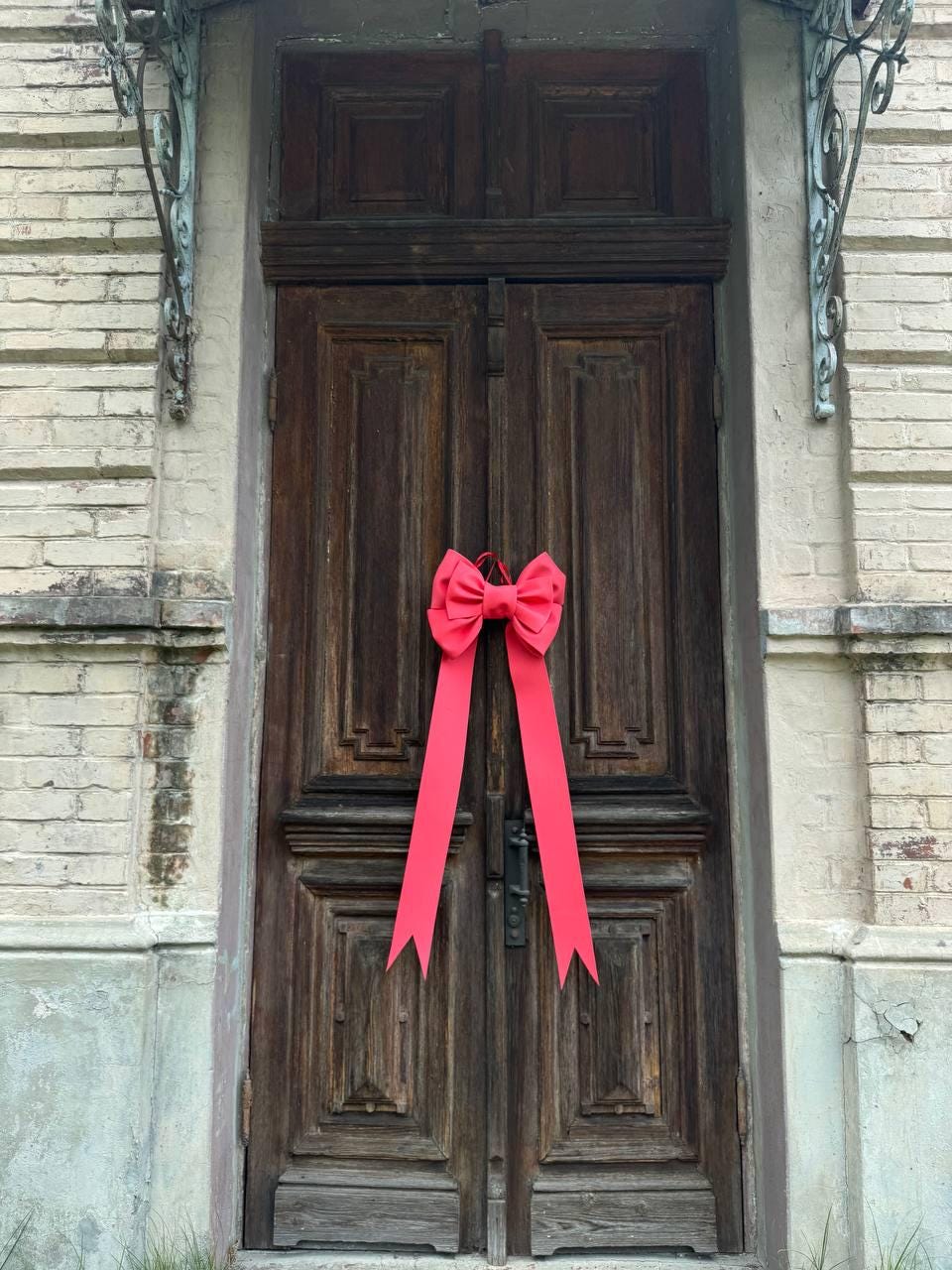 May include: A dark wooden door with a bright red bow. The door has a classic design with rectangular panels and a metal handle. The bow is centered on the door and has long, flowing ribbons.