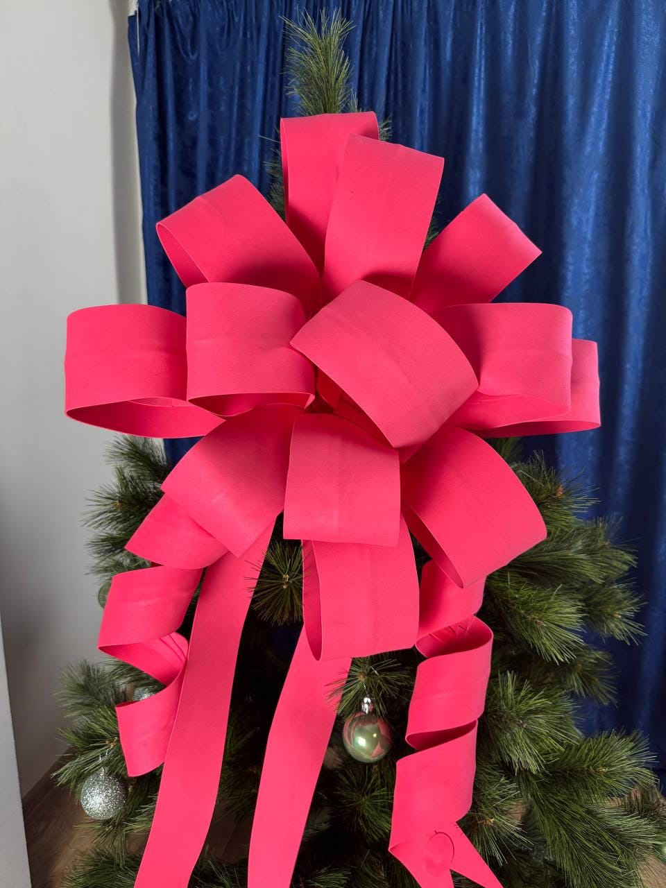 May include: A vibrant pink Christmas tree bow adorns a green artificial tree. The bow is made of wide, folded ribbon, with long, curling streamers. Silver and green ornaments are visible among the tree's needles. The background features blue curtains.