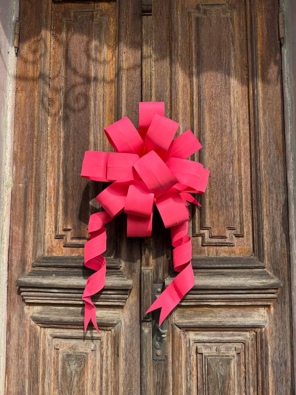 May include: A bright pink bow adorns a weathered wooden door. The bow's ribbons cascade downwards, adding a festive touch. The door features vertical panels and a vintage handle, creating a rustic aesthetic.