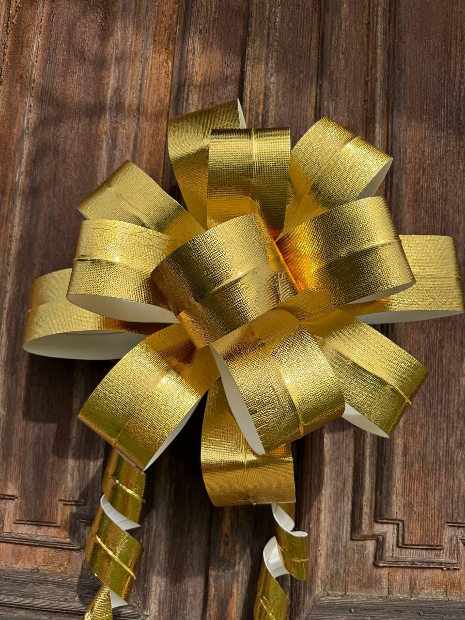 May include: A large, decorative gold bow with a textured surface. The bow is made of wide, metallic gold ribbon and has several loops and streamers. The bow is against a dark brown wooden background.