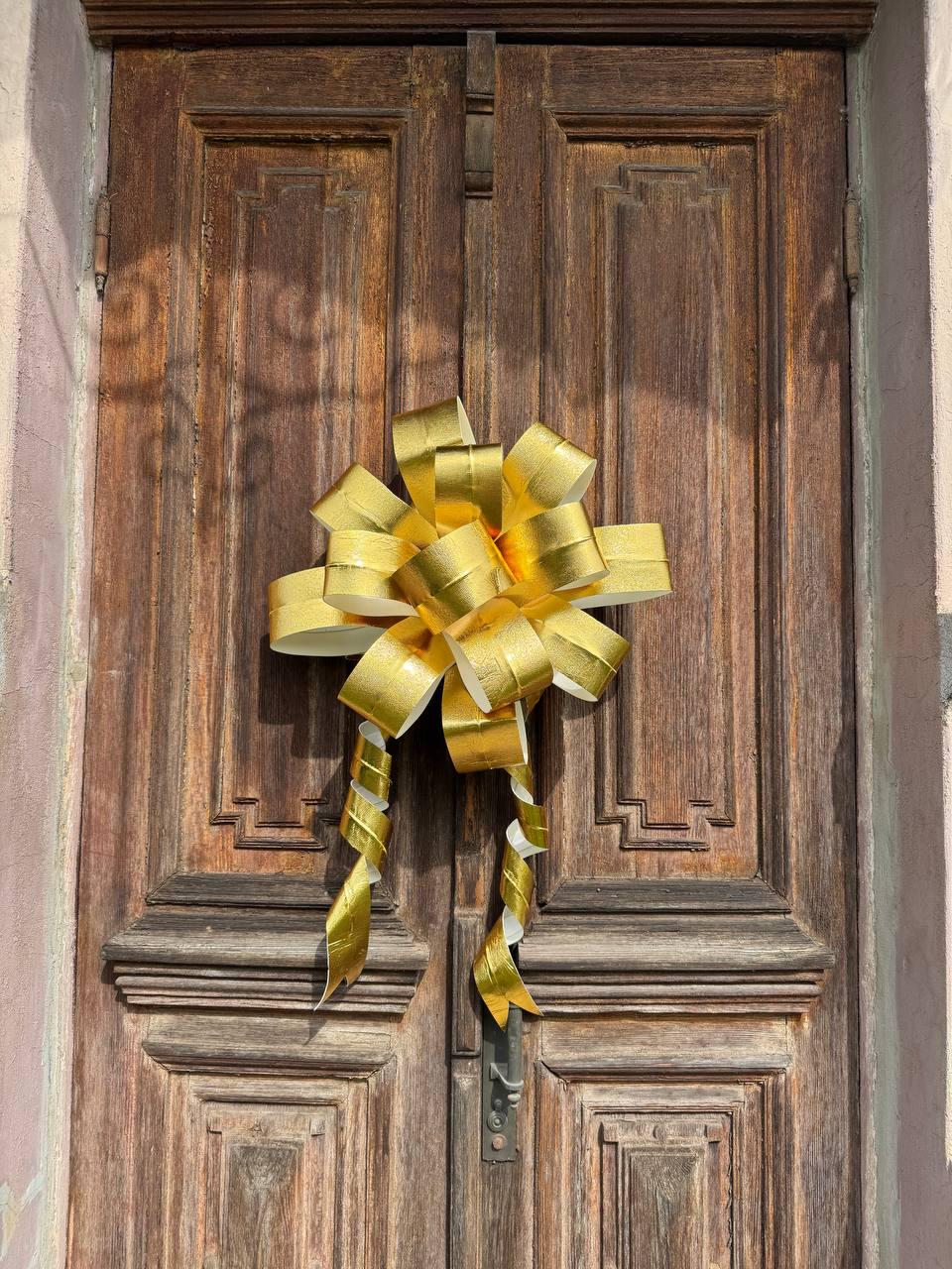 May include: A large, gold-colored bow adorns a weathered, wooden door. The bow is made of shiny, metallic material with long, curling streamers. The door has a dark brown finish with visible wood grain and decorative panels, suggesting a vintage or rustic style.