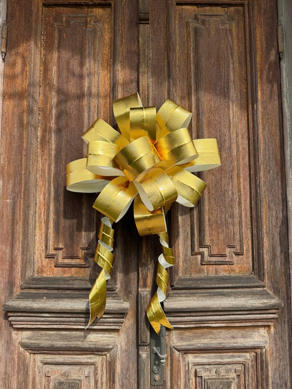 May include: A large, gold-colored decorative bow with spiraling streamers is affixed to a dark brown wooden door. The bow is made of shiny, metallic material, and the door has a detailed, paneled design. The bow is likely used for decoration.
