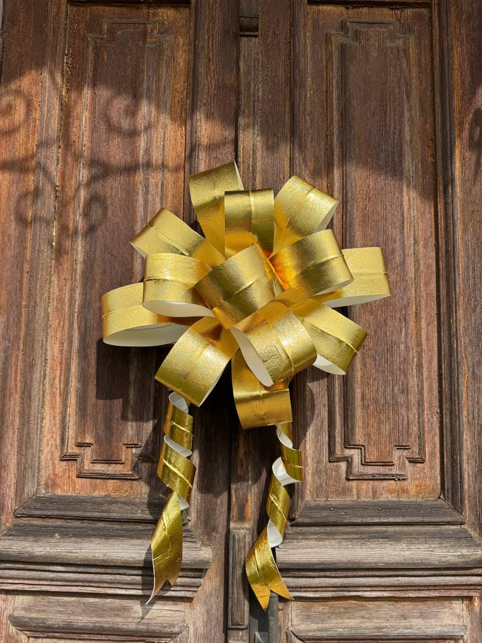 May include: A large, gold-colored bow with long, curled streamers is affixed to a dark brown wooden door. The bow is made of shiny, metallic material, and the door has vertical panels and decorative carvings.