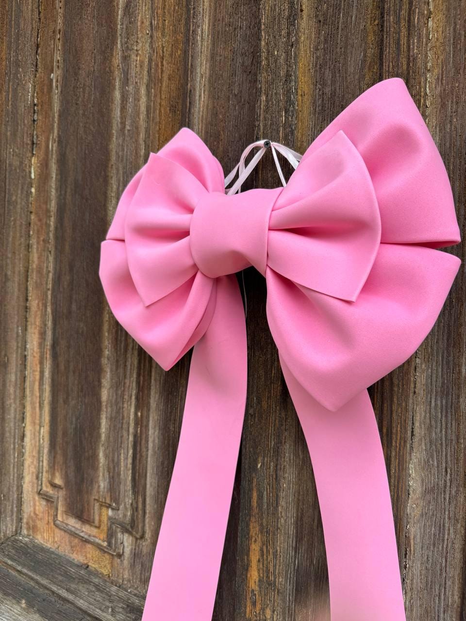May include: A large, bright pink bow with long, flowing ribbons. The bow is made of a smooth, possibly satin-like material. It is hanging against a rustic, wooden background, creating a contrast of textures and colors.